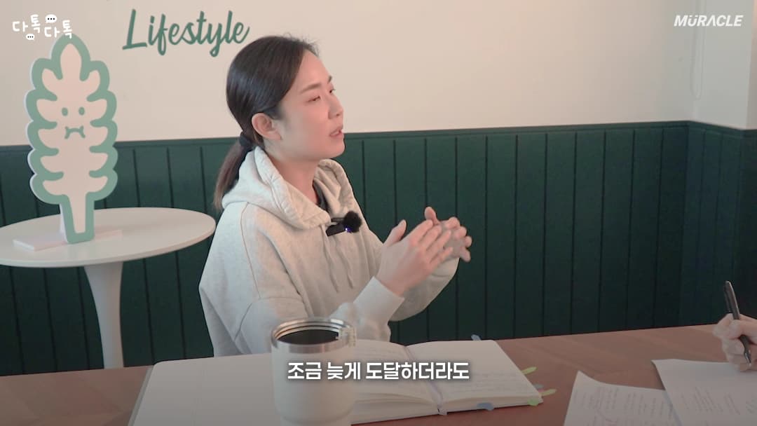 밝은 스튜디오에서 후드티를 입은 여성이 테이블에 앉아 손동작을 하며 대화하고 있다.