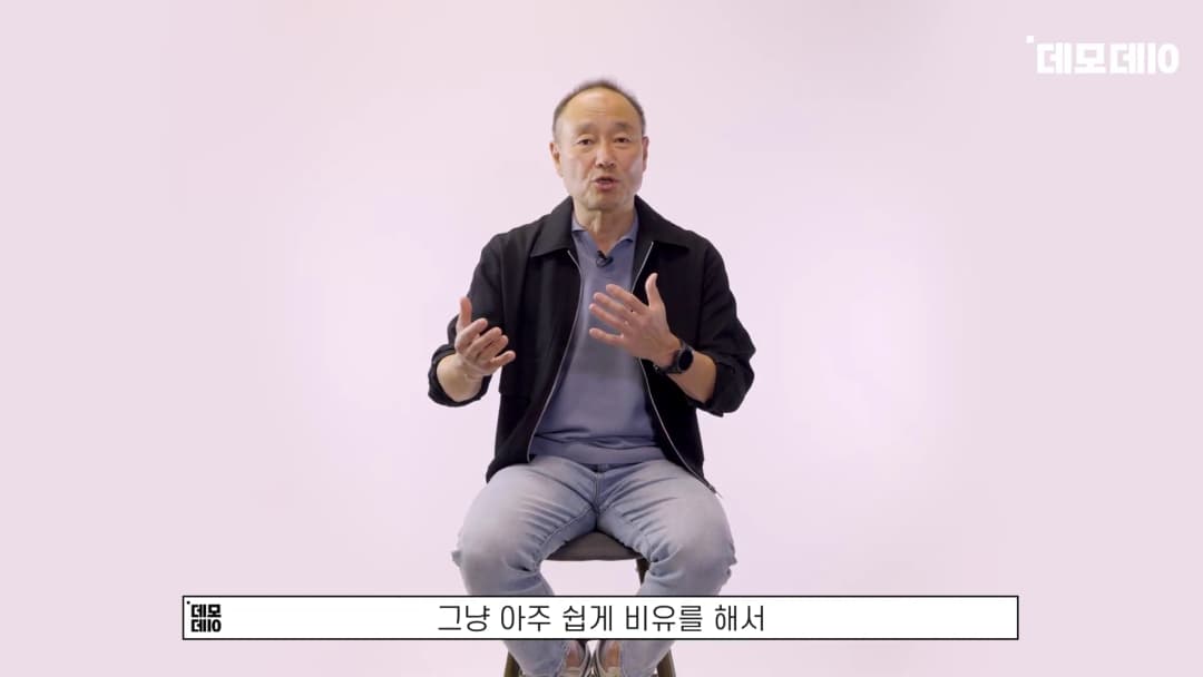 흰 배경의 스튜디오에서 의자에 앉아 손짓하며 이야기하는 중년 남성