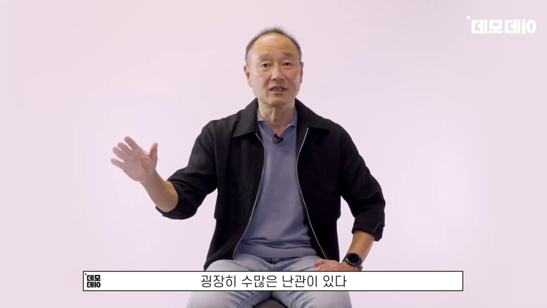 밝은 배경 앞에서 검은 재킷을 입은 중년 남성이 손을 들어 설명하고 있으며, 하단에는 자막이 표시되어 있다.