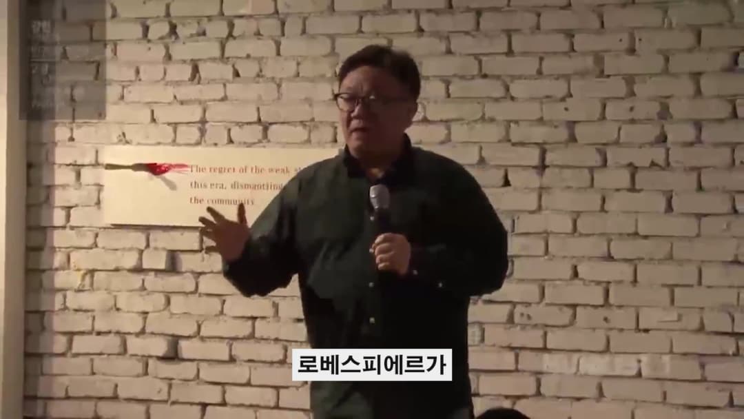 흰 벽돌 배경 앞에서 마이크를 들고 강연하는 중년 남성