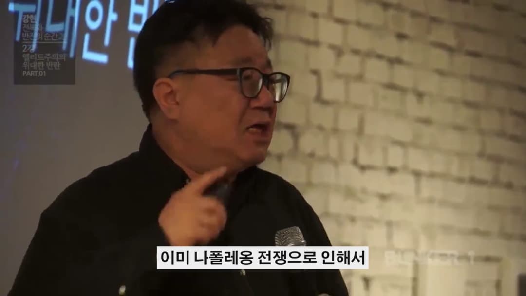 안경을 쓴 중년 남성이 강연장에서 마이크를 들고 열정적으로 이야기하고 있다.