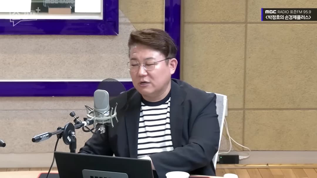 안경을 쓴 남성이 라디오 스튜디오에서 마이크 앞에 앉아 이야기를 나누고 있다.