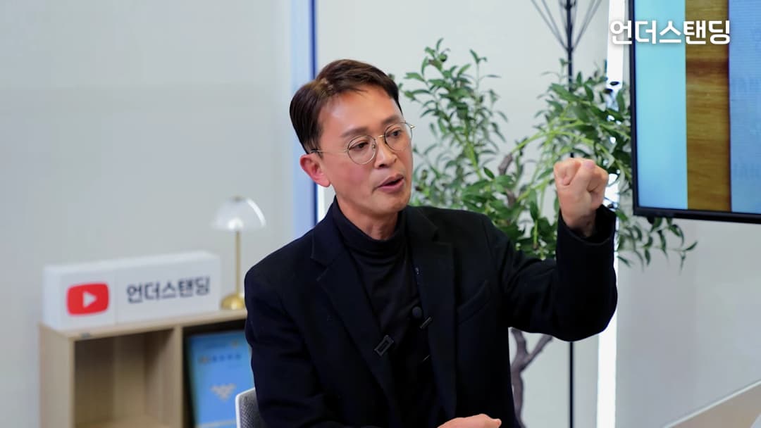 안경을 쓴 남성이 스튜디오에서 손을 들어 보이며 열정적으로 이야기하고 있다.