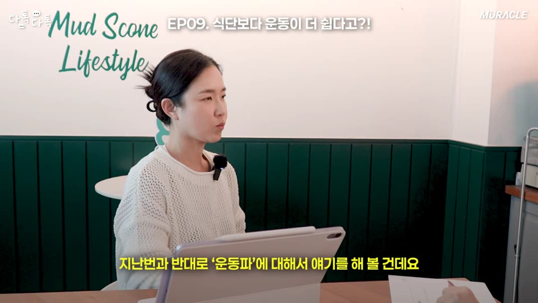 흰색 니트를 입은 여성이 테이블 앞에 앉아 이야기를 나누고 있는 스튜디오 인터뷰 장면