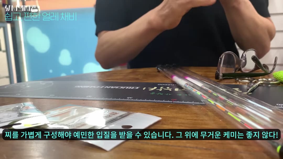 나무 테이블 위에 낚시찌와 도구들이 놓여 있고, 한 사람이 손으로 채비를 만지고 있는 모습