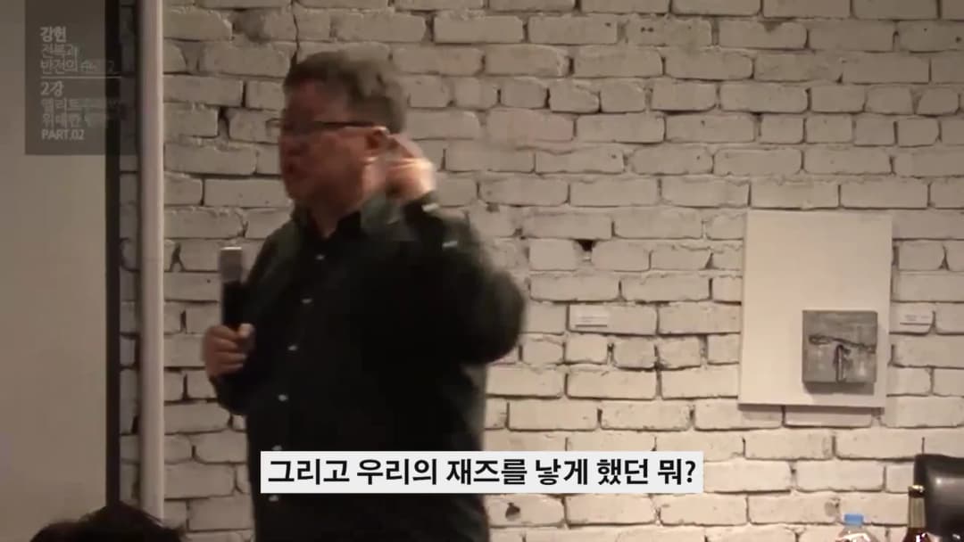 흰 벽돌 벽을 배경으로 마이크를 들고 강연하는 중년 남성