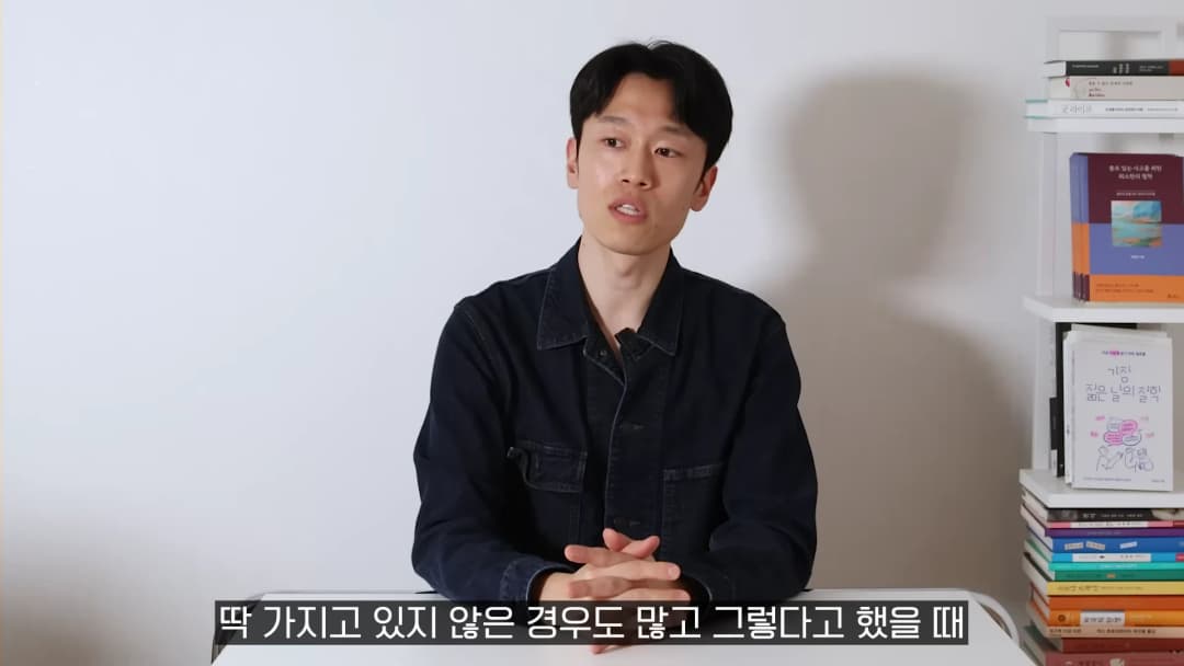 흰 벽을 배경으로 책장이 놓인 방에서 검은 셔츠를 입은 남성이 앉아 이야기를 나누고 있다.